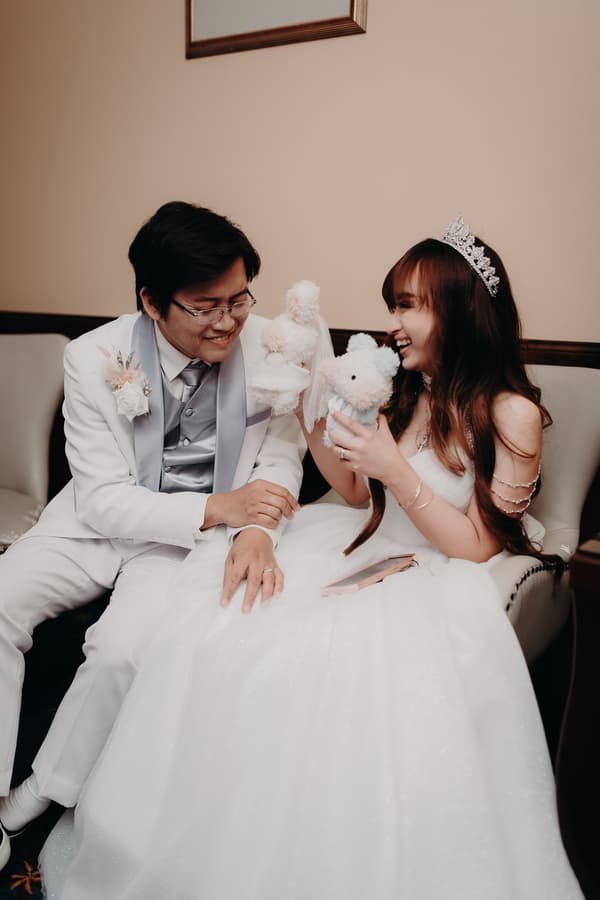 Timothy and Henny sit together on a couch at Royal on the Park during the reception, holding and playing with plush toys while dressed in wedding attire.