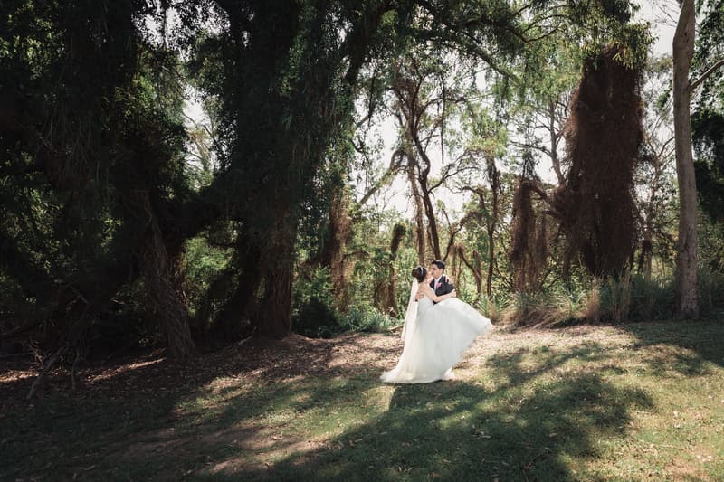 The bride and groom pose alone outdoors surrounded by large trees and greenery.