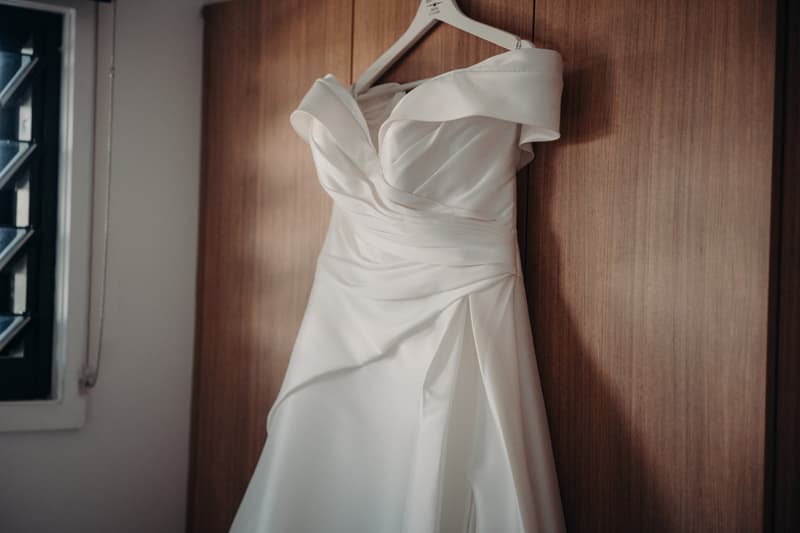 A white wedding dress hanging on a wooden door inside a room with a window nearby.