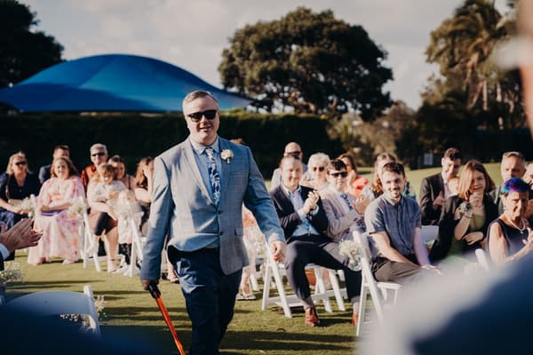 The groom Dale, wearing sunglasses and a light gray suit jacket with a floral tie, walks down the aisle at Sandstone Point Hotel — Rustic Arbour while guests seated on white chairs applaud.