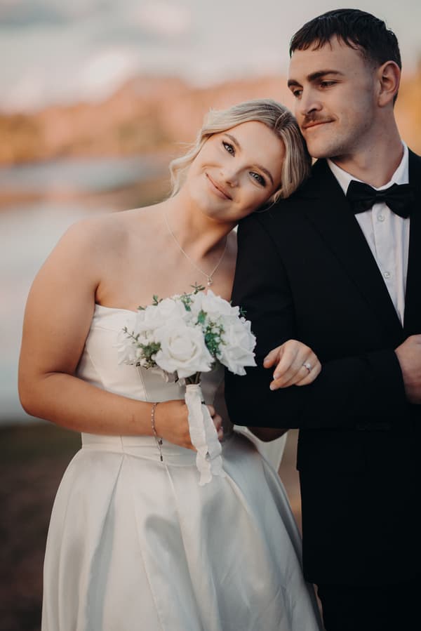 Libby, the bride, in a strapless white wedding gown holding a bouquet of white flowers, leans her head on Kyle, the groom's shoulder, who is dressed in a black tuxedo with a bow tie, posing outdoors at The Tides during their couple portraits session.