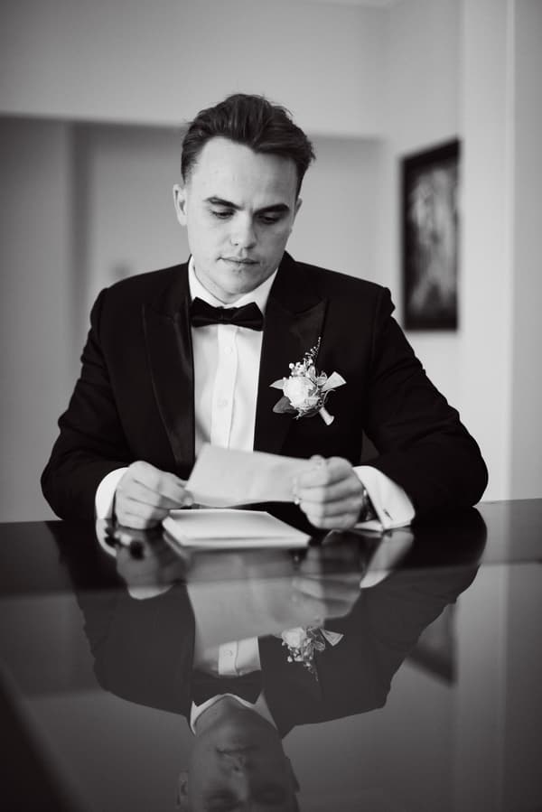 James, the groom, dressed in a tuxedo with a boutonniere, sits at a reflective table reading a piece of paper indoors.