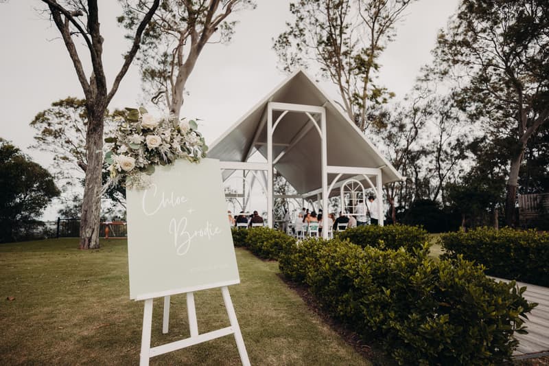 Wedding ceremony taking place under a white pavilion at Sandstone Point Hotel with guests seated and a floral welcome sign displaying the names Chloe and Brodie in the foreground.