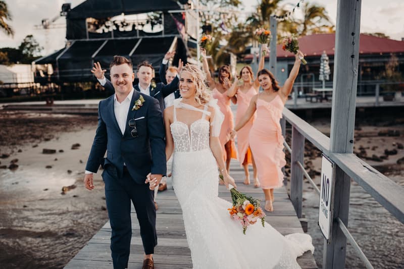 Bride Krystal in a white wedding gown and groom Brandon in a navy suit walk hand in hand on a wooden jetty at Sandstone Point Hotel, followed by bridesmaids in peach dresses holding bouquets.