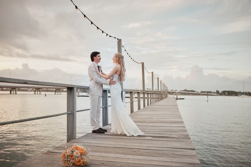 Morgan and Joel stand facing each other on a wooden pier at Sandstone Point Hotel, with string lights overhead and water surrounding the pier. A bridal bouquet lies on the pier near them.