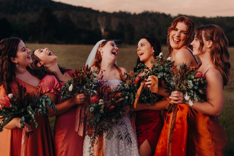 The bride Lilly in a white wedding dress and veil stands outdoors at Yabbaloumba Retreat with five bridesmaids dressed in various shades of orange and red dresses, all holding bouquets and laughing together.