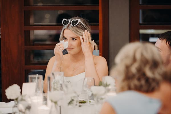 The bride Libby wearing heart-shaped sunglasses on her head wipes her eye with a tissue while seated at a table during the reception in The Pandanus Room at The Tides.