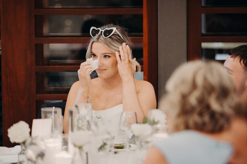 The bride Libby wearing heart-shaped sunglasses on her head wipes her eye with a tissue while seated at a table during the reception in The Pandanus Room at The Tides.