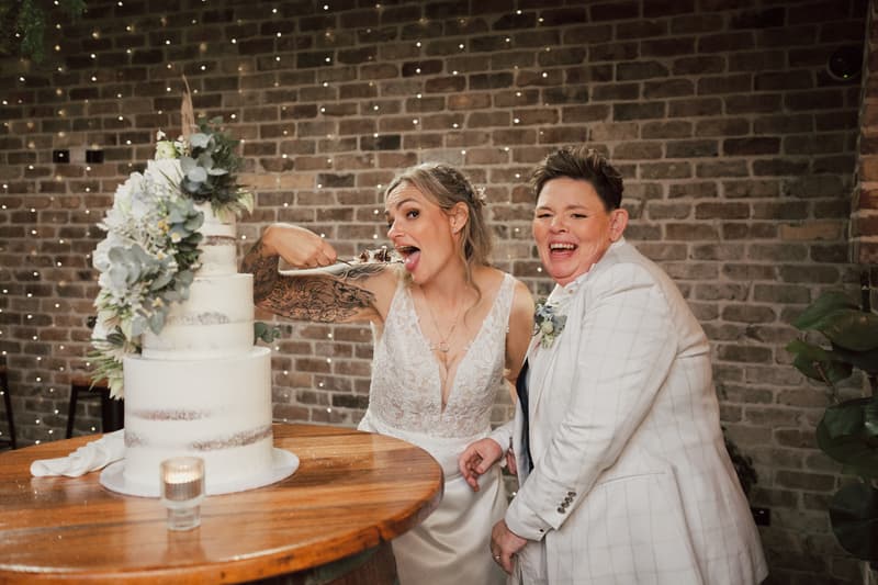 Brides Mandy and Selina pose together at the reception stage at Sandstone Point Hotel — The Cellar, with Mandy playfully holding a piece of wedding cake near her mouth beside a three-tier white wedding cake decorated with flowers.