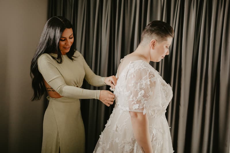 A woman in a light green dress is fastening the back of the bride's white lace wedding gown in front of dark curtains.