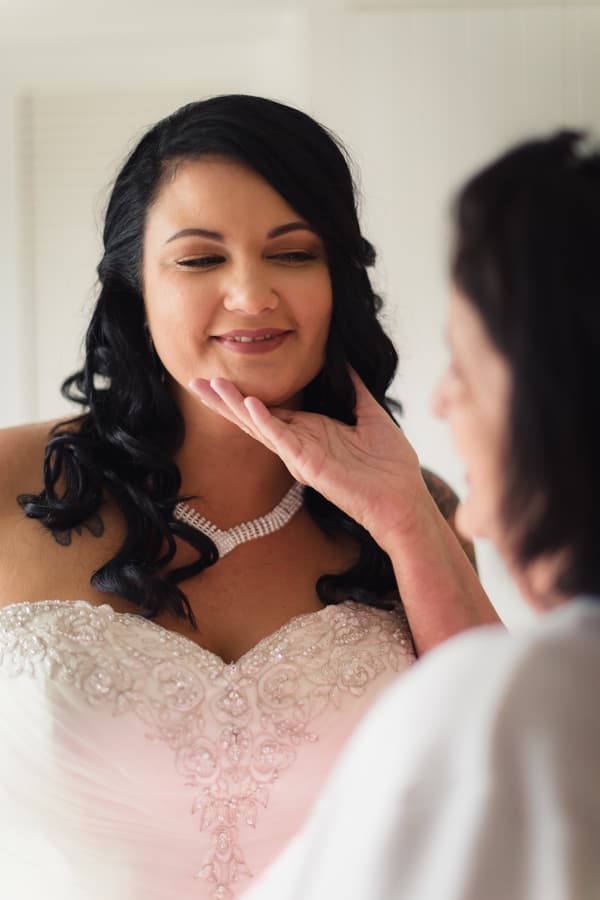 The bride Kelly in her wedding dress with detailed beadwork and a necklace, having her chin gently touched by an older woman, possibly the mother of the bride, at Novotel Sunshine Coast Resort.