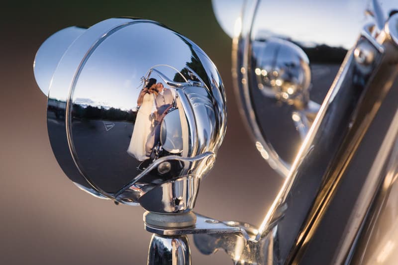 The bride Kelly and groom Shane are reflected kissing in the chrome headlight of a vintage car at Novotel Sunshine Coast Resort during their wedding day.