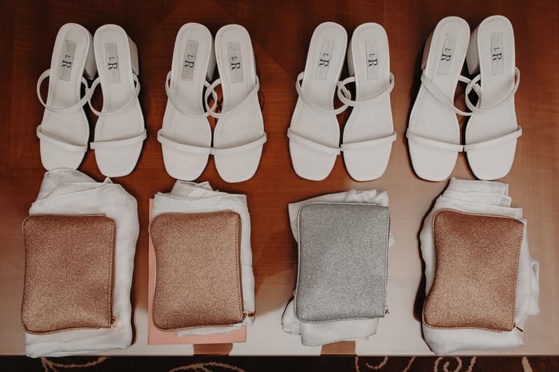 Four pairs of white heeled sandals arranged in a row on a wooden surface, each pair placed above folded white fabric and a small glittery clutch purse in rose gold or silver.