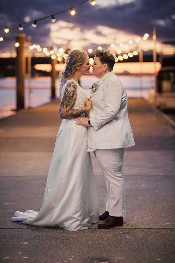 Mandy in a white wedding gown and Selina in a light-colored suit stand close together on a pier at sunset with string lights overhead at Sandstone Point Hotel.