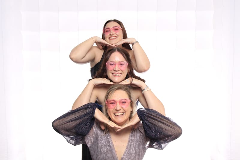 Three women posing playfully with pink sunglasses, hands under their chins.