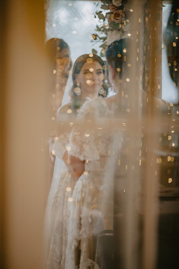 The bride in a lace wedding dress and veil is seen through a reflective surface with bokeh lights, looking at her reflection.