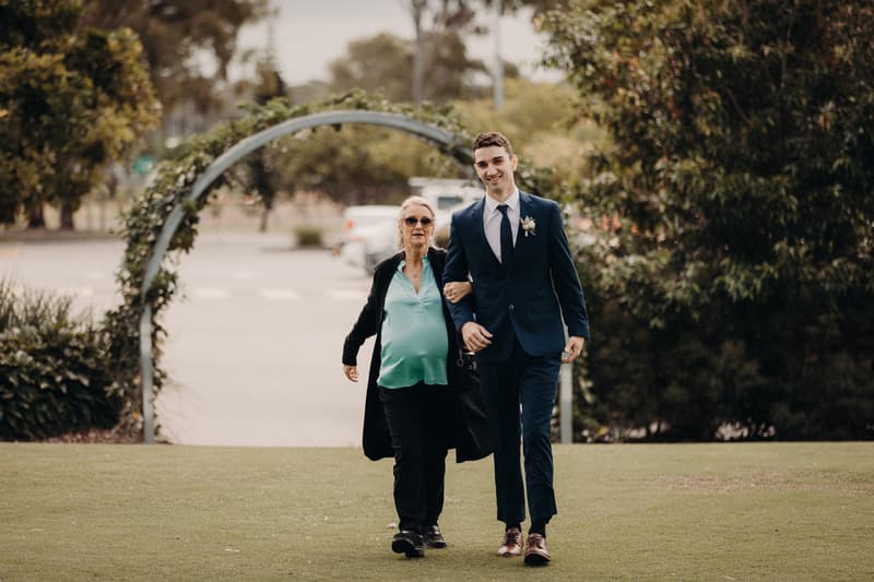 The groom Cameron walks arm-in-arm with an older woman across a grassy area at Sandstone Point Hotel — Pavilion, with a decorative arch and trees in the background.