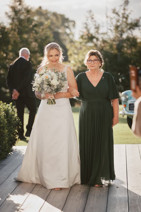 The bride Mandy walks arm-in-arm with an older woman, likely her mother, along a wooden pathway at Sandstone Point Hotel — The Pavilion during the ceremony stage. Mandy holds a bouquet of white and green flowers and wears a white wedding gown, while the older woman wears a dark green dress. A man in a suit and a vintage car are visible in the background.