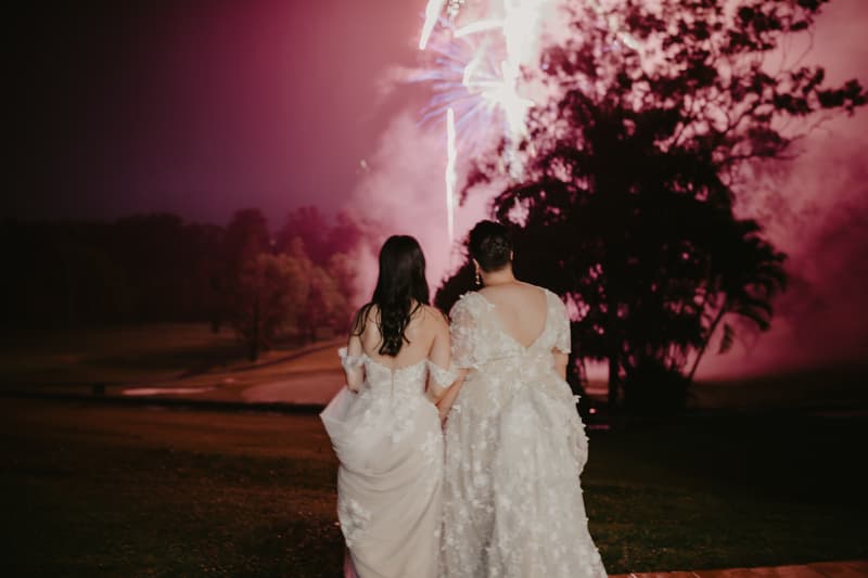 Two brides in white wedding dresses stand side by side outdoors at night watching fireworks in the sky.