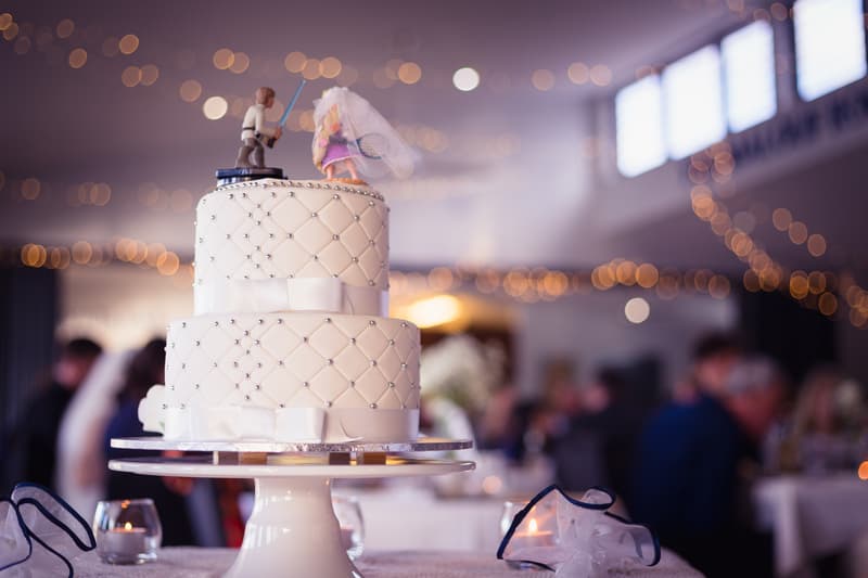 Two-tiered white wedding cake with quilted pattern and silver bead accents topped with bride and groom figurines, displayed on a table at the reception in The Malouf Room, Toowong Rowing Club.
