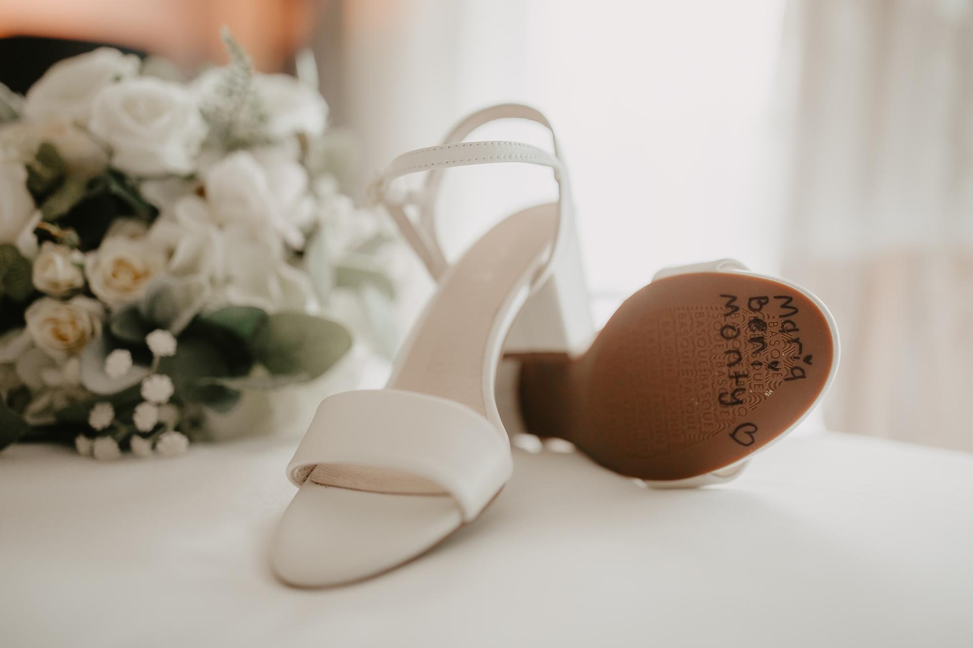 Close-up of a pair of white bridal shoes on a white surface with a bouquet of white flowers in the background. The sole of one shoe has handwritten names and hearts.