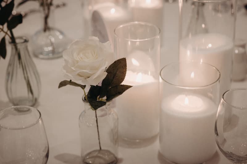 Close-up of a white rose in a small glass vase surrounded by lit white candles in glass holders on a table at The Tides — The Pandanus Room.