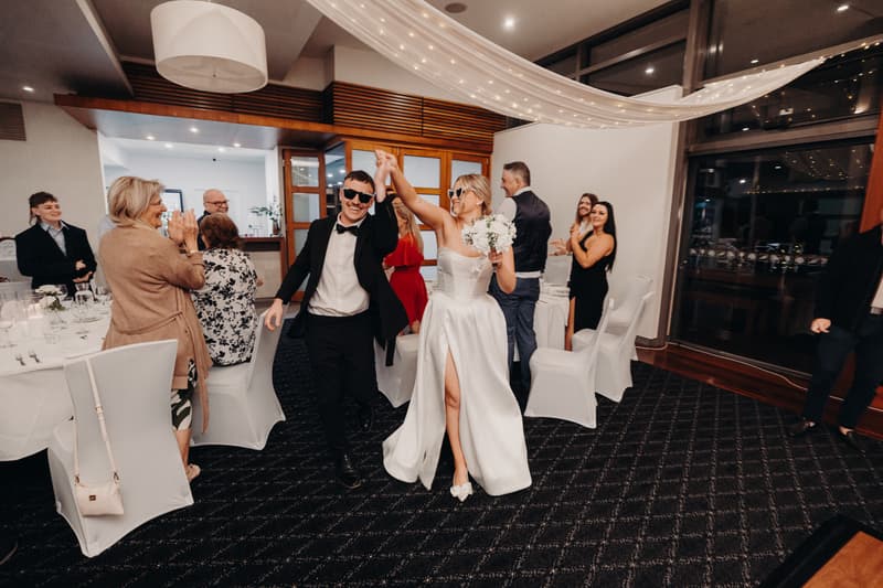 Bride Libby and groom Kyle wearing sunglasses hold hands and walk through The Pandanus Room at The Tides reception, while guests seated at tables applaud.