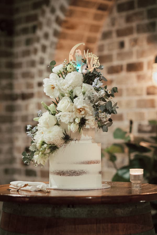 Two-tiered wedding cake decorated with white flowers and a 'Mrs & Mrs' topper on a wooden barrel table at Sandstone Point Hotel — The Cellar.