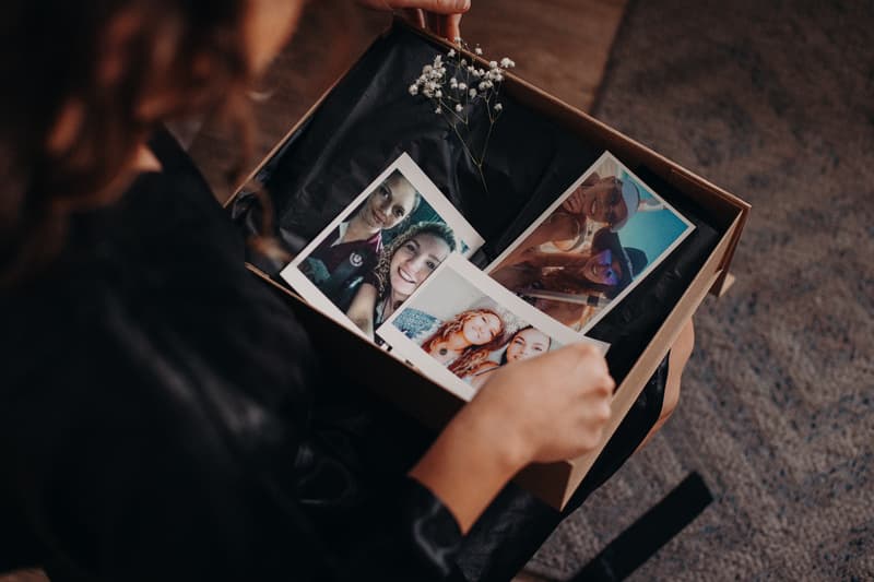 The bride looks at a box containing three printed photos of friends and a small sprig of baby's breath flower.