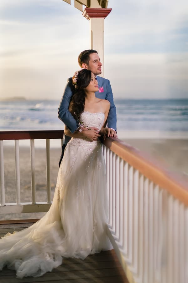 The bride Wing in a strapless white wedding gown and the groom Jason in a blue suit with a pink pocket square stand together on a wooden balcony overlooking the ocean at Bilinga Beach Weddings.