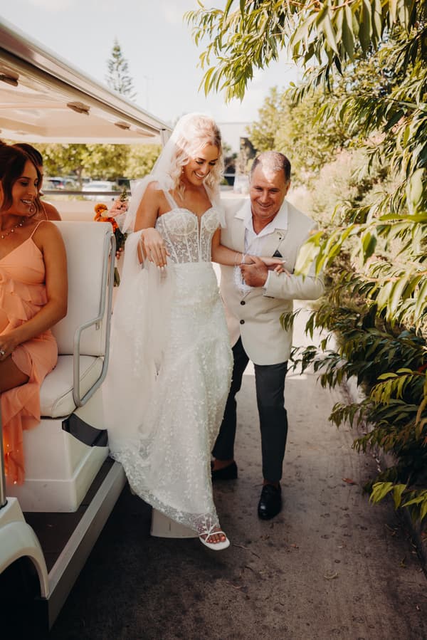 The bride Krystal, wearing a white wedding dress and veil, is stepping down from a vehicle with assistance from an older man, likely her father, at Sandstone Point Hotel — Pavilion. A bridesmaid in a peach dress is seated inside the vehicle watching.