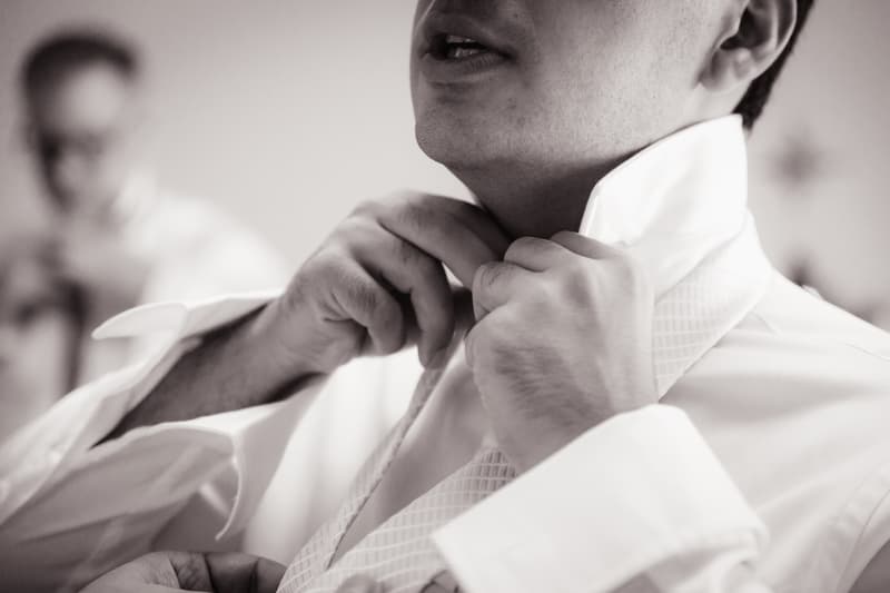 The groom adjusts his tie while getting ready, with another person blurred in the background.