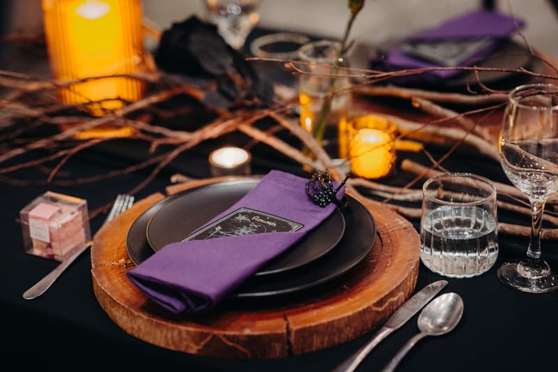 Close-up of a wedding reception table setting at Ocean View Estates — Function Room featuring a wooden charger plate, black plates, a purple napkin with a decorative black crown, glassware, cutlery, candles, and rustic twig centerpiece.