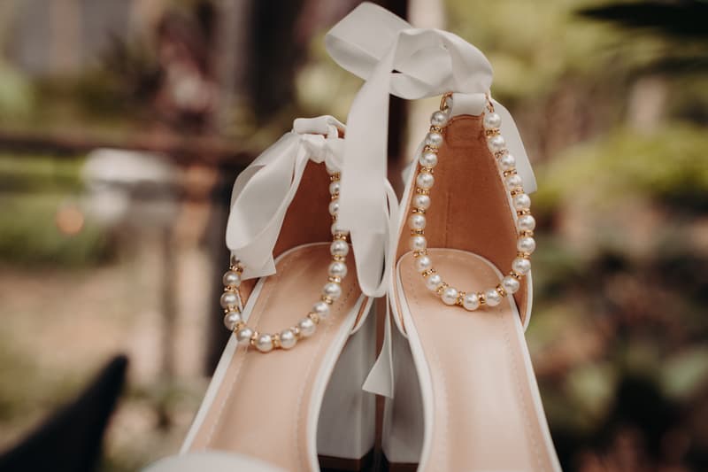 Close-up of a pair of white bridal shoes with pearl and gold bead ankle straps, photographed outdoors.