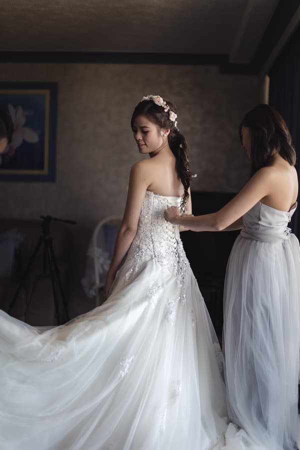 The bride Wing is having the back of her wedding dress laced up by a bridesmaid in a softly lit room.