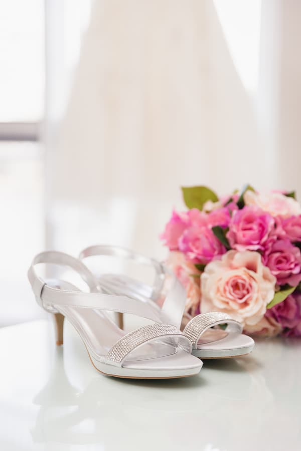 The bride's silver high-heeled shoes are placed on a reflective surface with a bouquet of pink and peach roses in the background at Novotel Sunshine Coast Resort.