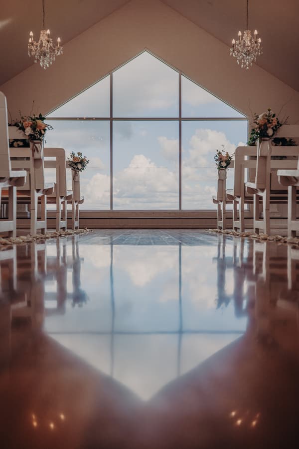 Empty chapel aisle at Tiffany's Maleny with floral decorations on white pews and large triangular window showing a cloudy sky.