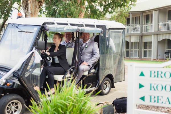 The groom Shane and two other men sit in a golf cart driven by a woman outside a building at Novotel Sunshine Coast Resort.