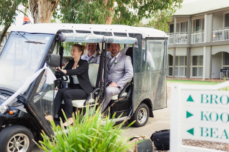 The groom Shane and two other men sit in a golf cart driven by a woman outside a building at Novotel Sunshine Coast Resort.