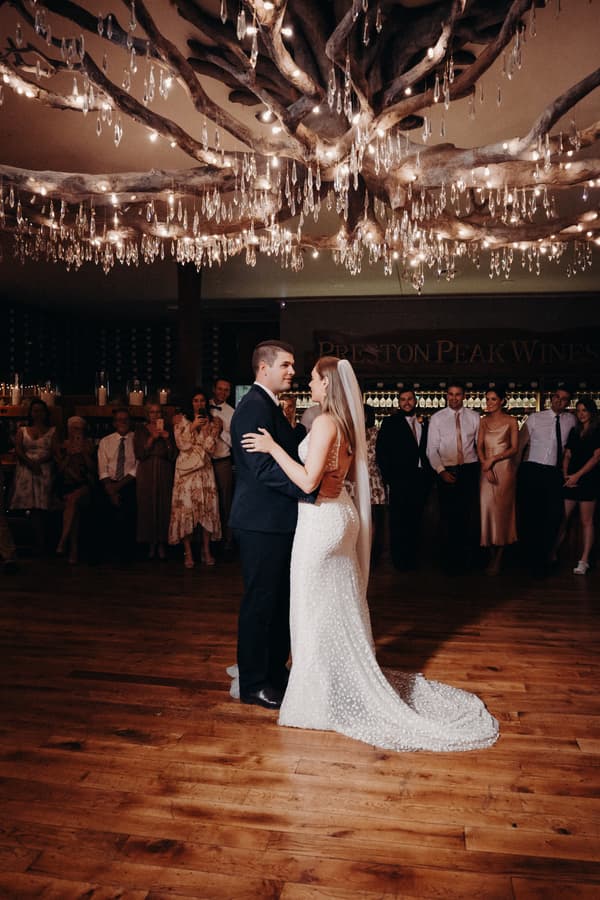 Bride Chelsea and groom Brent share a dance on the wooden floor at Preston Peak Winery Functions Centre, surrounded by guests watching in the background under a large decorative chandelier.