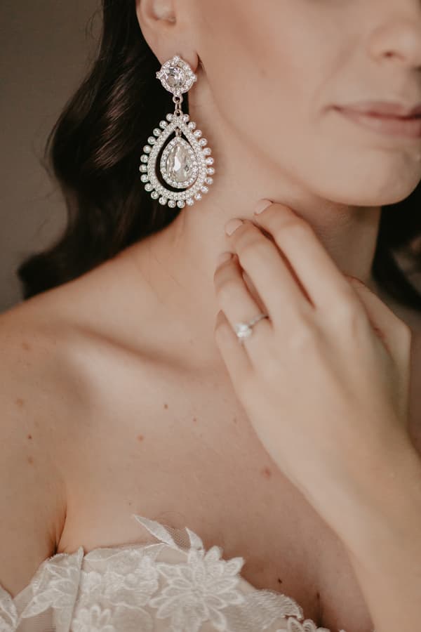 Close-up of a bride wearing a strapless lace wedding dress, a large teardrop-shaped crystal earring, and an engagement ring on her left hand touching her neck.
