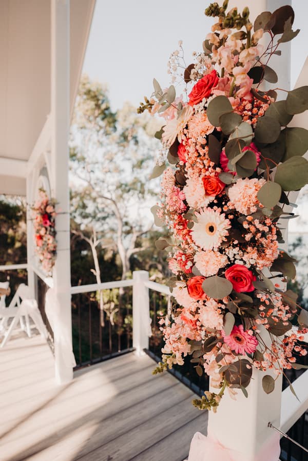 Floral arrangements with pink and red flowers decorate white pillars at the Pavilion of Sandstone Point Hotel for a wedding ceremony.