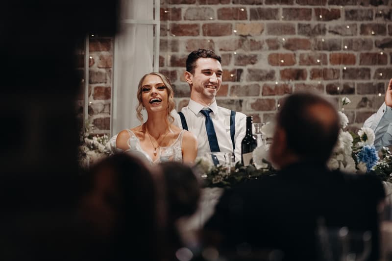 Courtney the bride and Cameron the groom sit at a decorated reception table at Sandstone Point Hotel — Cellar, smiling and facing guests.