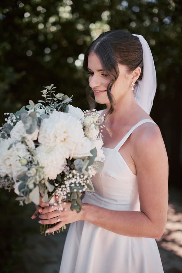 The bride Ashleigh in her wedding dress holding a large bouquet of white flowers and greenery, standing outdoors with a blurred natural background.