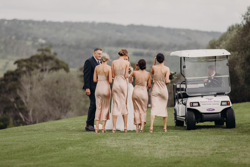 The bride Chelsea, groom Brent, and four bridesmaids walk on grass near a golf cart at Preston Peak Winery Functions Centre with a scenic hillside background.