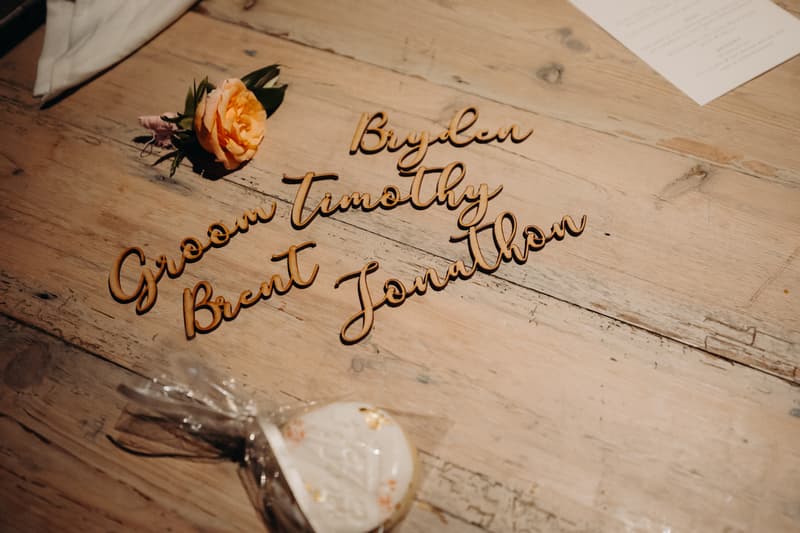 Wooden name cutouts including 'Groom Brent' and other names arranged on a wooden table at Preston Peak Winery Functions Centre, alongside a peach rose boutonniere and a wrapped cookie favor.