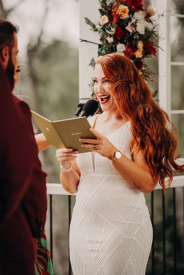 The bride with long red hair reads from a booklet into a microphone during the ceremony at Sandstone Point Hotel — The Pavilion, facing the groom who is partially visible.