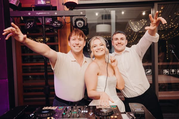 The bride Libby wearing headphones stands between two men behind DJ equipment at The Tides — The Pandanus Room during the reception stage.