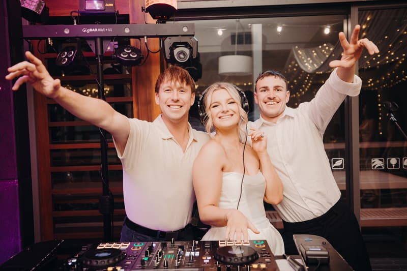 The bride Libby wearing headphones stands between two men behind DJ equipment at The Tides — The Pandanus Room during the reception stage.
