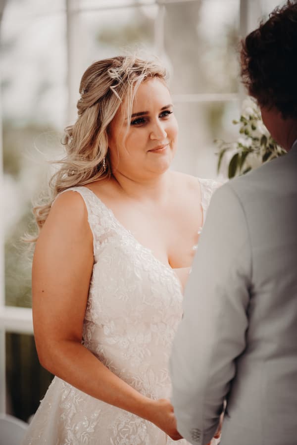 The bride Chloe in a white lace wedding dress faces the groom Brodie, holding hands during the ceremony at Sandstone Point Hotel — Pavilion.